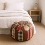 Colorful patchwork round leather pouf ottoman placed in a soft minimalist bedroom beside a natural wood nightstand.