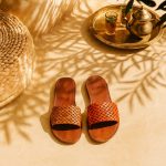 Pair of hand-woven orange Leather Slides resting in dappled palm-leaf shade next to a brass mint-tea tray.