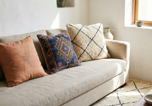 An assortment of handmade Moroccan pillow covers, including cactus silk and wool Kilim styles with geometric patterns, layered on a sofa in a bright, modern room.
