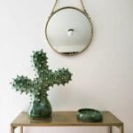 Green ceramic cactus sculpture with spiky texture beneath round brass mirror on gold console table