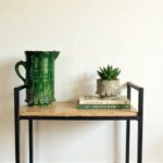 Artisan green ceramic pitcher on industrial metal stand with stacked books and small succulent planter