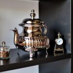 Handcrafted ribbed silver teapot ornate feet and domed lid on dark shelf beside a vintage brass clock