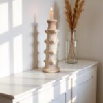 White ceramic tower candlestick with organic sculptural ridges displayed on cabinet beside glass vase with dried grasses