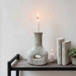 Rustic ceramic candle holder with lit taper, sculptural gray pottery on wooden shelf with books