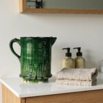 Green glazed ceramic pitcher styled in minimalist bathroom setting with folded towels and wood mirror