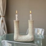 Speckled ceramic double candleholder with two lit white candles on glass table in neutral room setting