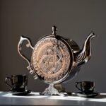 Geometric engraving on decorative silver teapot display with ball finial lid and splayed feet near teacups