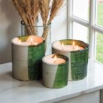 Artisanal ceramic candle collection with mottled green metallic finish burning by window with wheat grass