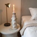 Tall white ceramic candlestick tower with tiered design holding burning candle on bedside table next to brass lamp