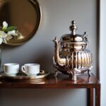 Handcrafted ribbed silver teapot ornate details on a wooden sideboard with teacups and a decorative mirror