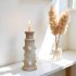 White ceramic candlestick tower with sculptural tiered design holding lit candle next to pampas grass in glass vase