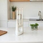White ceramic candle holder with three candles on kitchen counter next to plants and cutting board