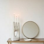 Modern ceramic candle holder with three lit candles on wooden bench beside round mirror and dish