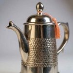 Close-up of handcrafted silver-plated brass coffee pot featuring hammered honeycomb texture and decorative orange tassel on lid