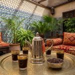 Silver-plated brass coffee pot with hammered finish displayed in traditional Moroccan riad courtyard with zellige tiles, brass tray, and Arabic coffee glasses