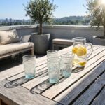 Sunlit white geometric tea glasses resting on a slatted wooden patio table, accompanied by a glass pitcher of fresh lemon water on a sunny rooftop terrace with a city view.