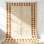 Handwoven wool rug with cream pile and terracotta border squares displayed vertically on white wall