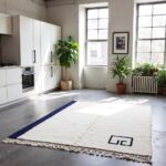Modern wool rug in industrial loft kitchen with cream base, navy diagonal border, and knotted fringe
