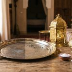 A still life photograph of freshly polished, shiny Moroccan brass items, including a teapot and tray, arranged on a table next to natural cleaning supplies like lemons, salt, and a soft cloth.