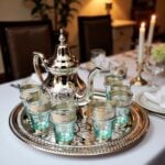 Silver engraved beldi tea set on a dark wood table showing a footed teapot and aqua glasses with metal bands