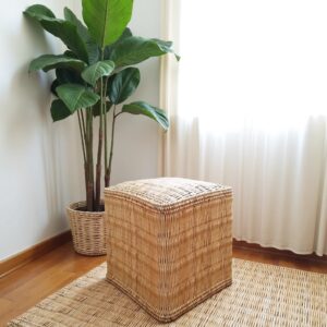Handwoven square Moroccan straw pouf beside a potted plant, creating a natural, minimalist corner on wooden flooring with matching straw mat.