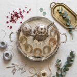 Top-down flat lay of a large moroccan tea set in silver, featuring a teapot, sugar bowl, and six glasses on a handled tray with eucalyptus and rose petal accents.