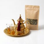 A traditional brass mini tea set for two including a golden teapot, tray, and glasses, placed next to a kraft paper bag filled with loose leaf green tea on a white surface.