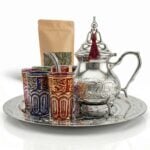 A complete moroccan mint tea set featuring an intricately engraved silver teapot with a red tassel, vibrant multicolored tea glasses with gold patterns, and a matching silver tray, displayed alongside a kraft paper pouch of loose leaf tea against a solid white background.