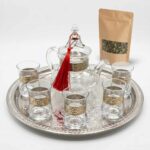 An elegant silver engraved glass tea set featuring a clear glass teapot decorated with a red tassel and an ornate metal band, accompanied by six matching glasses. The set is beautifully arranged on an intricately engraved silver tray alongside a kraft paper pouch of loose leaf green tea against a crisp white background.