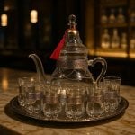 Elegant silver engraved glass tea set with clear teapot and 6 glasses on ornate metal tray, dark ambiance