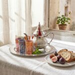 Lifestyle shot of a moroccan mint tea set on a linen-covered table with fresh figs and bread, situated by a sunlit window.