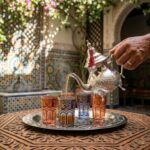 Steam rises as tea is poured into a moroccan mint tea set in a lush riad courtyard with mosaic zellige tilework in the background.