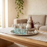 Steaming hot silver engraved beldi tea set resting gracefully on a light beige linen runner over a wooden coffee table, situated in a bright, modern living room setting.