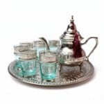 Silver engraved beldi tea set featuring an authentic Moroccan teapot adorned with a burgundy tassel, six blue-tinted glasses with intricate engraved silver metal sleeves, and a coordinating round serving tray, isolated on a bright white background.