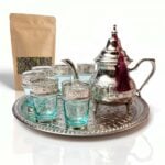 Complete silver engraved beldi tea set arranged neatly beside a kraft paper pouch of loose leaf green tea on a clean white surface, showcasing the traditional brewing presentation.