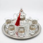 Studio product photography of a silver engraved glass tea set featuring a glass teapot and six glasses with intricate silver bands on a matching engraved tray.