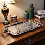 Silver serving tray with scrolled handles styled on wooden console beside gold lamp