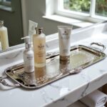 Rectangular silver tray with arabesque border holding skincare bottles on white marble vanity