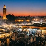 A wide-angle view of Jemaa el-Fna square in Marrakech at dusk, showing smoky food stalls, crowds of people, and the Koutoubia Mosque minaret illuminated against the evening sky.
