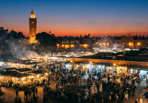 A wide-angle view of Jemaa el-Fna square in Marrakech at dusk, showing smoky food stalls, crowds of people, and the Koutoubia Mosque minaret illuminated against the evening sky.