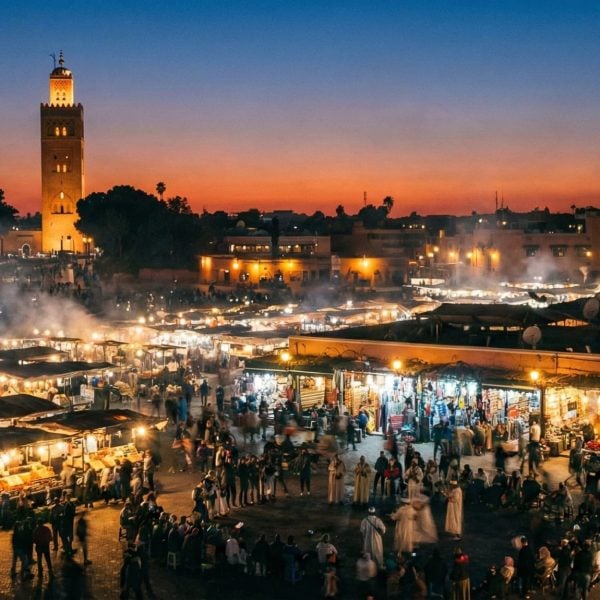A wide-angle view of Jemaa el-Fna square in Marrakech at dusk, showing smoky food stalls, crowds of people, and the Koutoubia Mosque minaret illuminated against the evening sky.