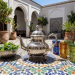 Hand-chased silver teapot wheat leaf design on blue zellige mosaic table in sunlit courtyard with tea glasses