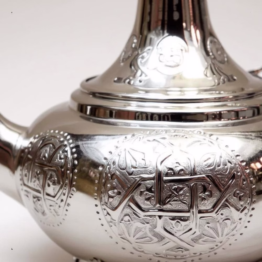 Close-up of engraving on a silver large teapot, showing intricate geometric and floral patterns with reflective metal finish.