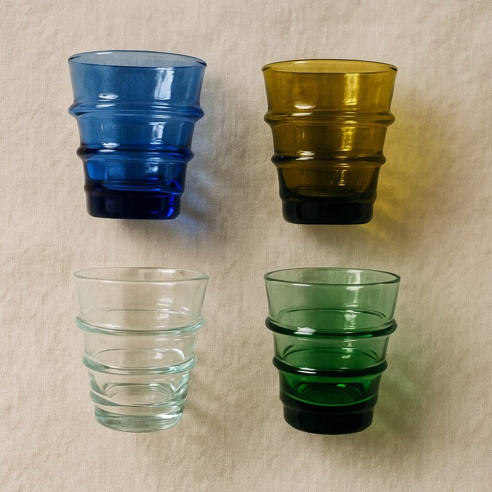 Flat-lay of four jewel-tone glass tumblers (blue, amber, clear, green) casting colored shadows on linen background.