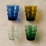 Flat-lay of four jewel-tone glass tumblers (blue, amber, clear, green) casting colored shadows on linen background.