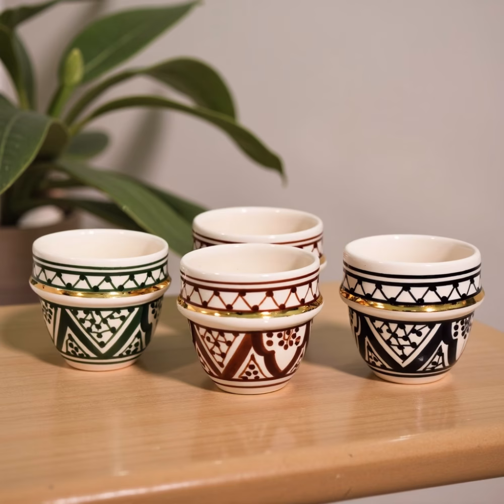 Four Moroccan espresso cups in green, amber, black, and blue geometric motifs with 24-k gold bands on wood shelf.