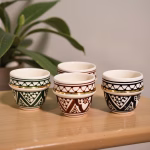 Four Moroccan espresso cups in green, amber, black, and blue geometric motifs with 24-k gold bands on wood shelf.