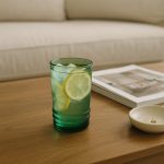Green tumbler of iced lemonade on wood table in cozy neutral living room.