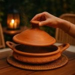Hand removing conical lid from terracotta tagine pottery revealing cooking chamber, traditional clay vessel design
