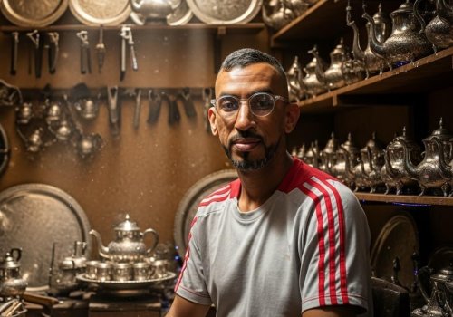 Kamal, a Moroccan silver-plated crafts artisan, looking confidently in front of shelves filled with traditional teapots and trays.